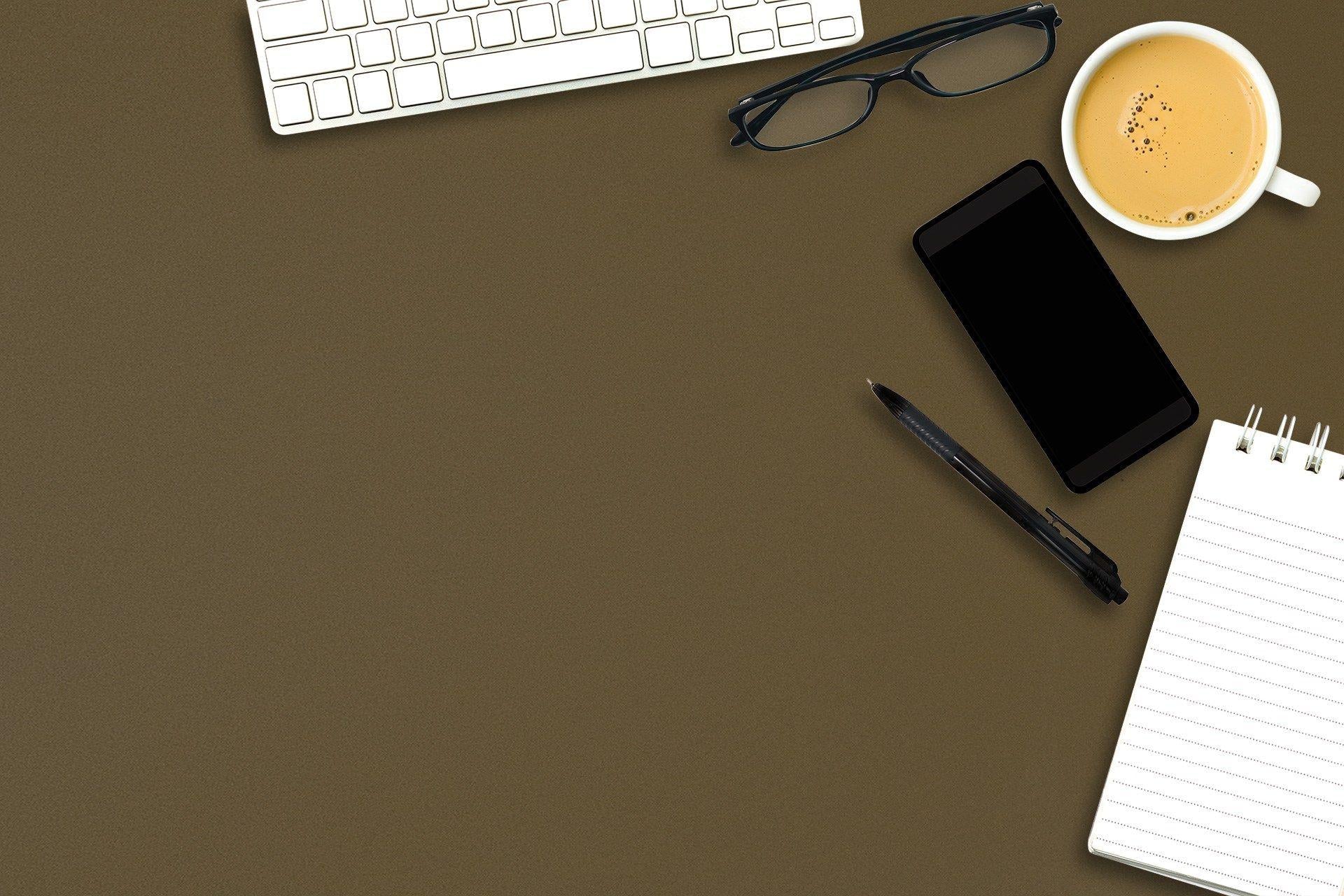 Brown suede laminate surface with office accessories including keyboard, glasses, phone, pen and notebook on top.