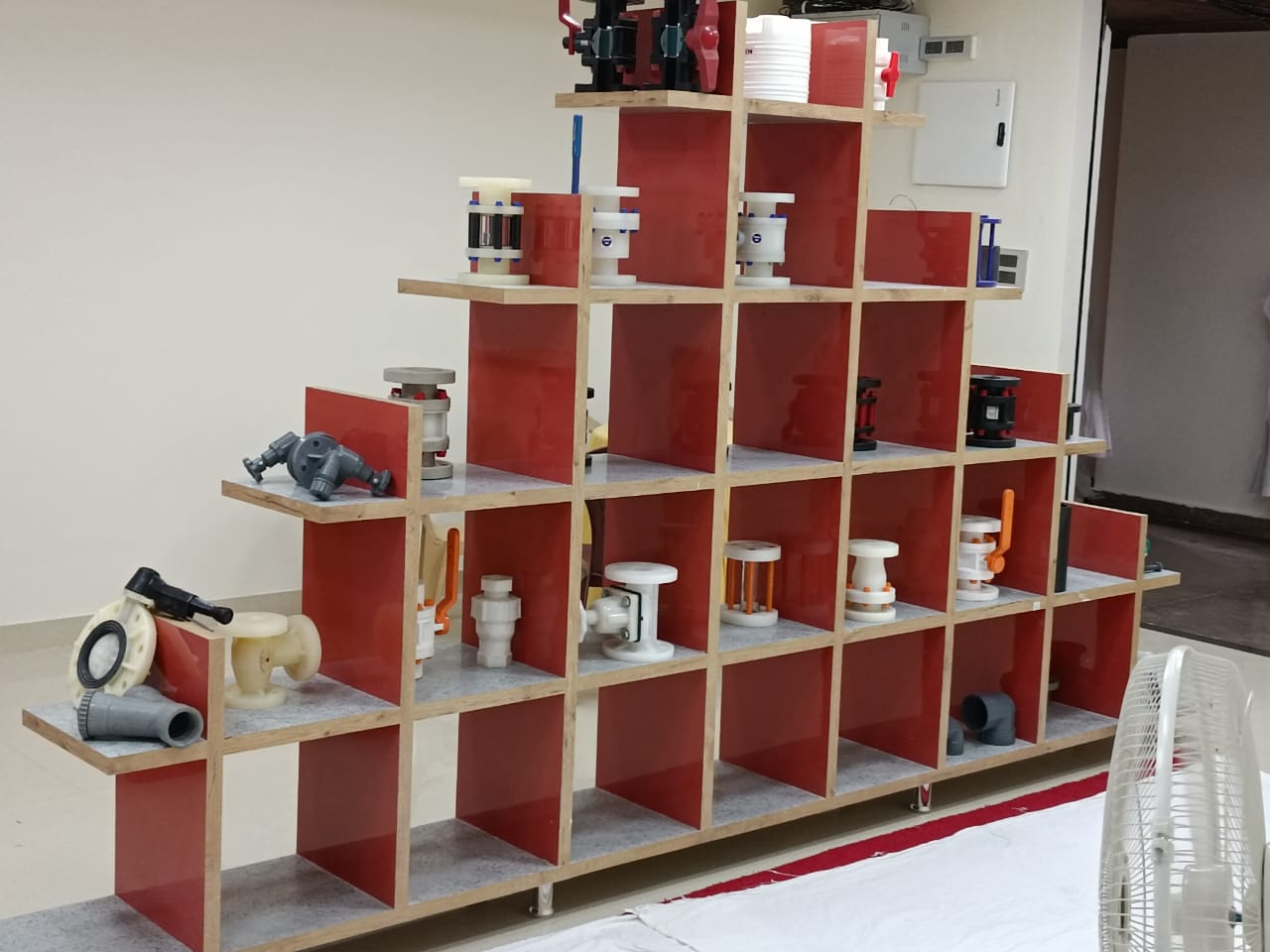Red laminate shelving unit displaying various plumbing parts and pipes in a room with white walls and dark floor.